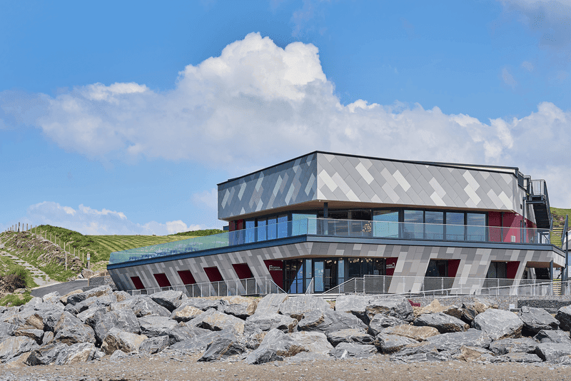 Edificio moderno arroccato sulla costa rocciosa, con tetto piatto e grandi finestre di vetro, in un cielo blu con nuvole sparse.